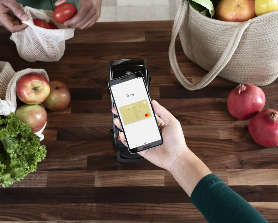 Ein Handy mit der Google Pay Funktion und integrierter TF Mastercard Gold wird über einen Tisch gehalten. Daneben liegen Lebensmittel-Einkäufe.