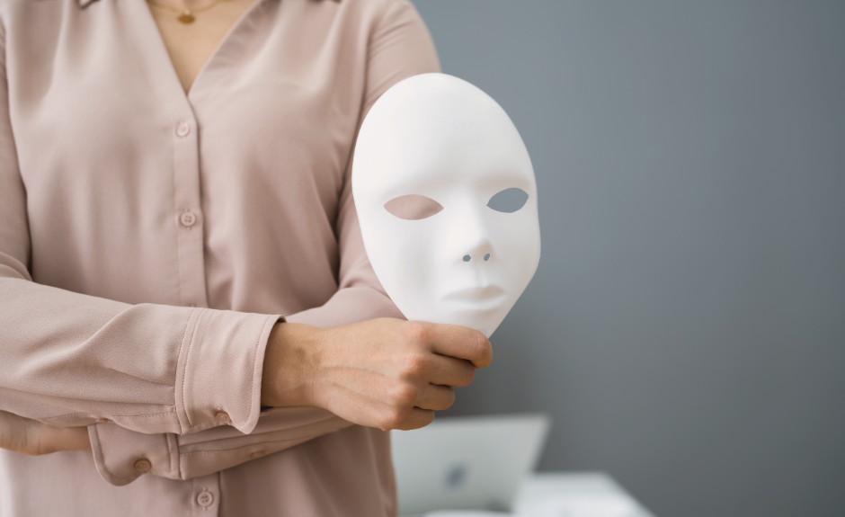 EIne Frau hält mit verschränkten Armen eine weiße Maske eines Gesichts in der Hand.