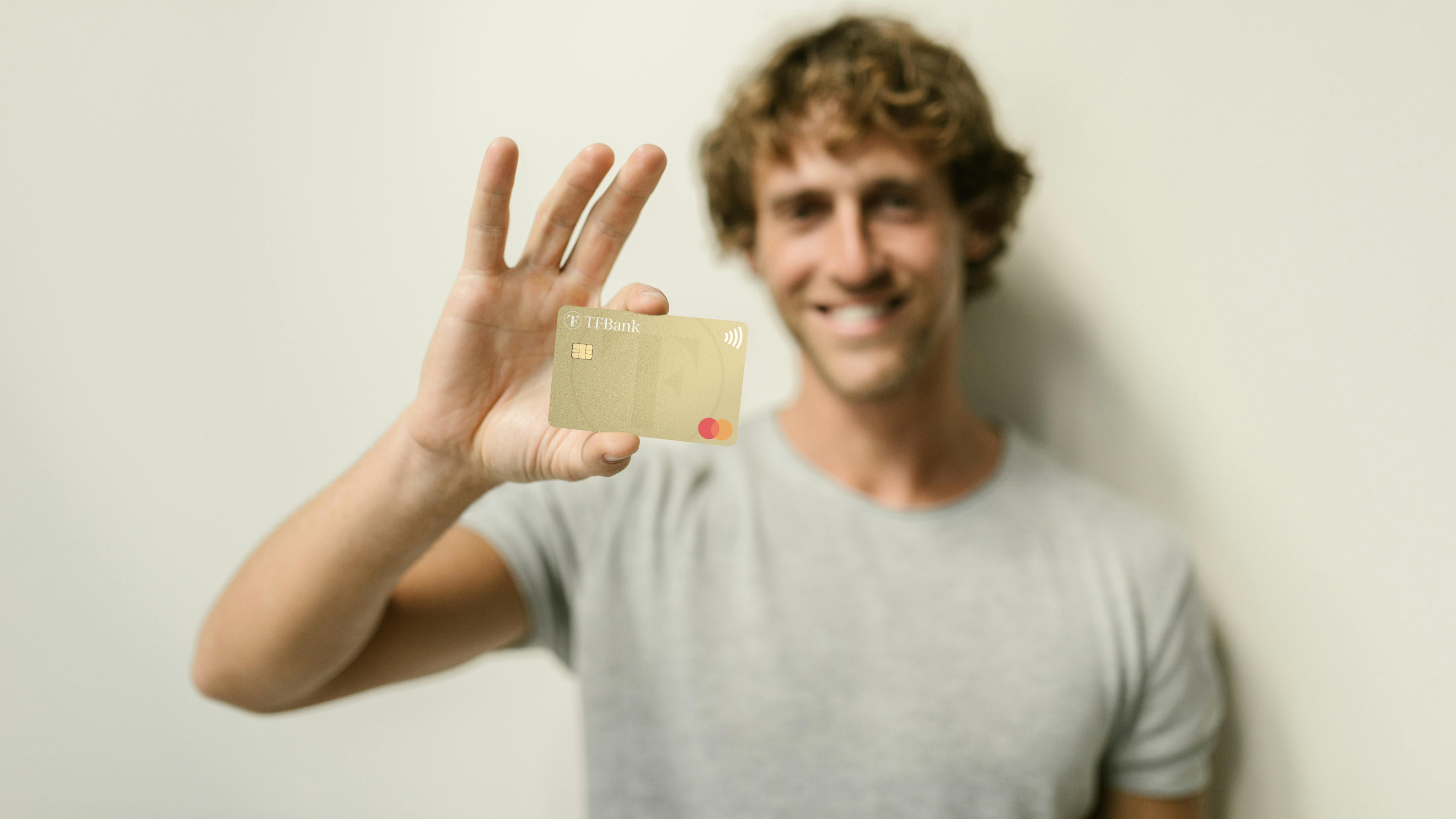 Ein Mann hält lächelnd die TF Mastercard Gold mit der rechten Hand in die Kamera. Fokus liegt auf der Karte. Der Mann ist leicht verschwommen.
