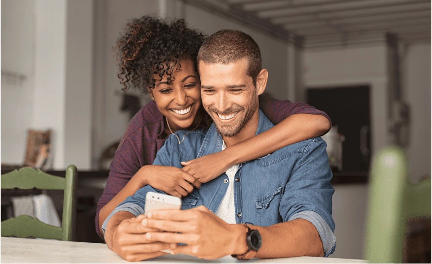 Man sitzt am Handy und eine Frau umarmt ihn von hinten. Sie grinsen beide.