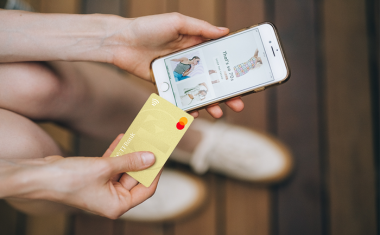 Es ist ein Iphone mit geöffnetem Onlineshop abgebildet. Daneben wird die TF Mastercard Gold in der Hand gehalten.