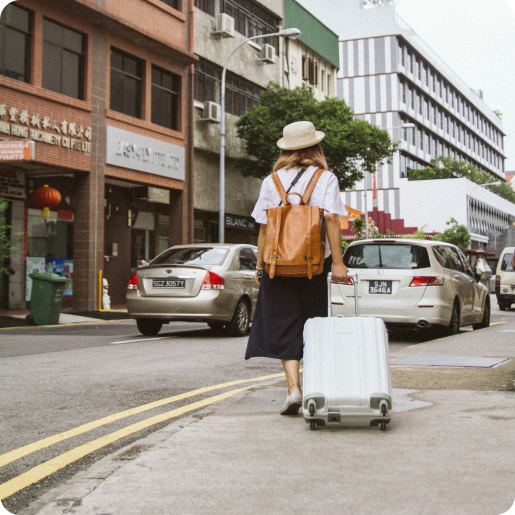 Eine Frau läuft auf dem Gehweg entlang einer Straße im Ausland. Man sieht sie von hinten mit einem Hut auf. Sie zieht einen Koffer hinter sich her.