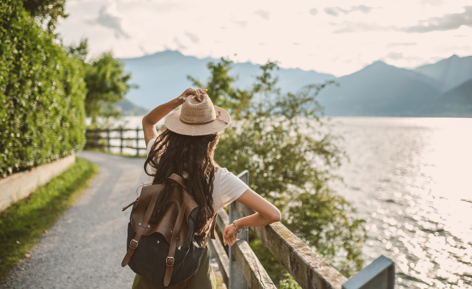 Eine Frau mit Rucksack steht mit dem Rücken zur Kamera und fasst sich auf ihren Strohhut. Sie lehnt an einem Geländer und schaut aufs Wasser.