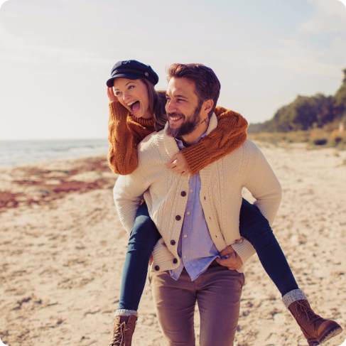 Ein Mann trägt eine Frau auf dem Rücken. Sie sind am Strand und beide lachen.