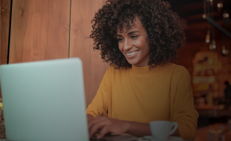 Frau sitzt am Tisch und tippt lächelnd etwas in ihren Laptop. Danben steht ein Kaffee.