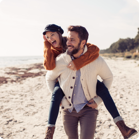 Ein Mann trägt eine Frau auf dem Rücken. Sie sind am Strand und beide lachen.